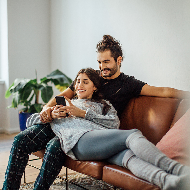 Smiling young woman using mobile phone while reclining on boyfriend sitting on sofa at home